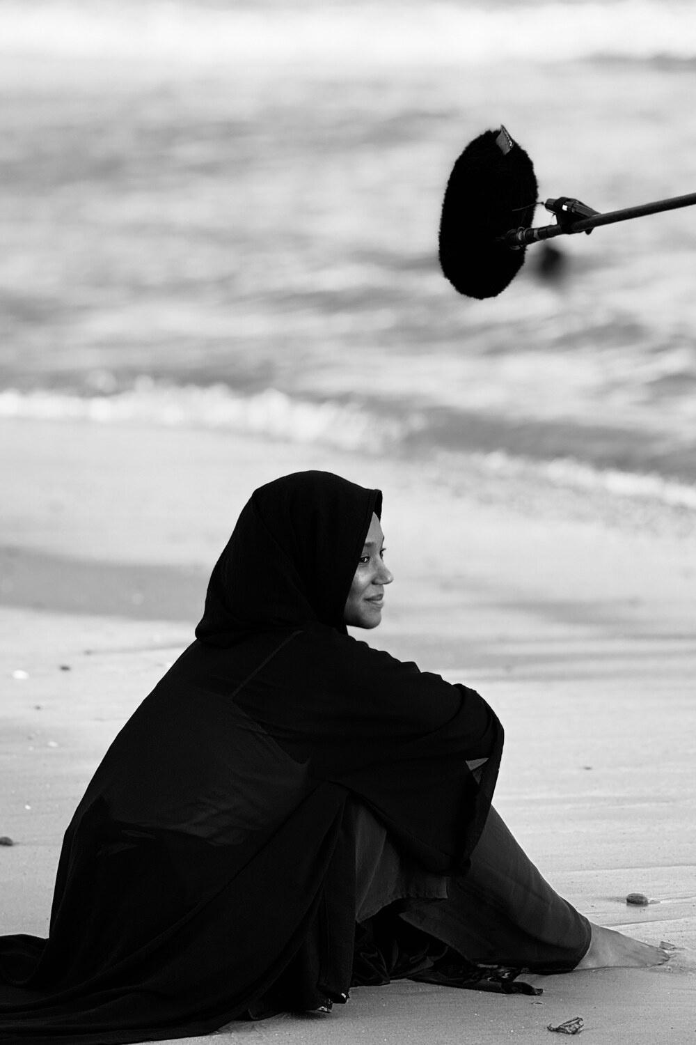 Fighters — photo de tournage en noir et blanc : Amy Lucia Diop assise sur le sable, drapée de noir, une perche de son au-dessus d'elle